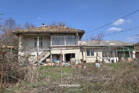 View of a two-storey house showing external stairs, an enclosed balcony, and surrounding dry grass and vegetation