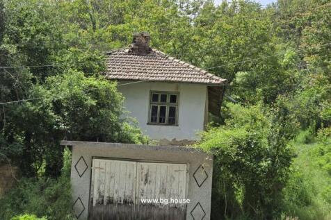 Close-up view of the old Bulgarian house showing the tiled roof, garage door, and surrounding green bushes.