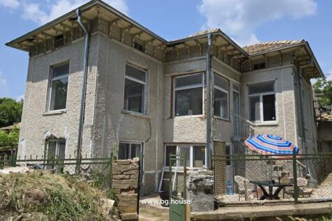 Side perspective of the Bulgarian house highlighting the textured walls, windows, and fenced yard with table and chairs under a striped umbrella