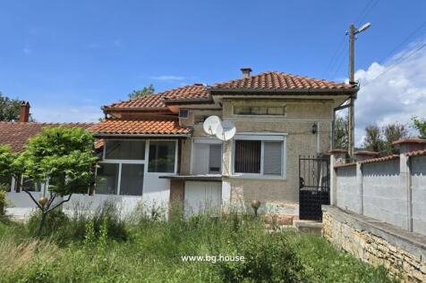 Exterior view of a Bulgarian house with terracotta tiled roof, satellite dishes, and a garden with green trees and grass.