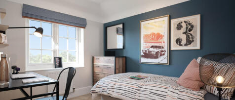 A cosy bedroom with a bed, colourful artwork on a blue wall, a wooden chest of drawers, and a desk w