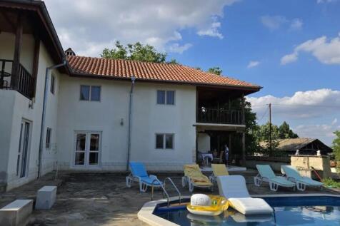 View of the white two-storey house with a tiled roof and a swimming pool surrounded by sun loungers on a stone patio.