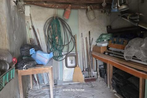 Interior view of a storage room featuring wooden beams, a sink, garden hoses, brooms, and various stored materials on shelves and floor.