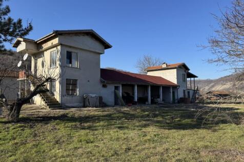 View of a two-story rural house with an attached single-story section and a grassy yard with leafless trees.