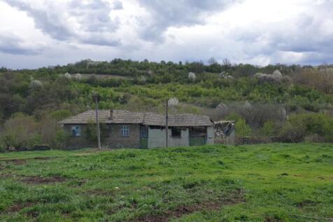 View of a traditional rural house with a tiled roof set against a backdrop of green hills and trees under a cloudy sky.
