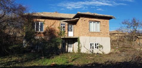 Front view of a two-storey rural Bulgarian house made of brick and stone with a tiled roof and surrounding trees.