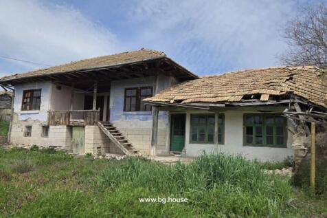 Exterior view of a traditional Bulgarian house showing stone basement, wooden balcony, tiled roof, and green doors and windows.