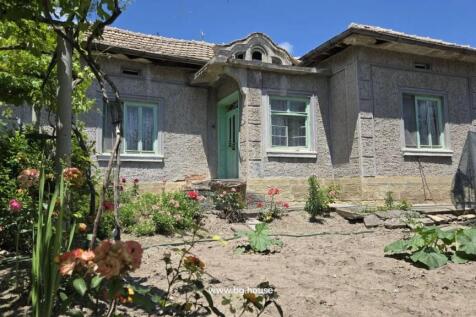 Close-up of the front entrance of a rural house featuring a green door and windows, with flowers and garden plants in the foreground.
