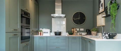 Modern green kitchen with built-in oven, microwave, black clock, and decorative plants on shelves at