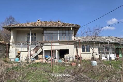 Front facade of a two-storey Bulgarian house showing external stairs and storage area beneath, with dry grass in the foreground