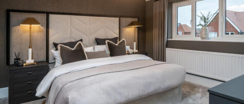 A cosy bedroom with a large bed, dark pillows, bedside lamps, a window with a view of neighbouring h