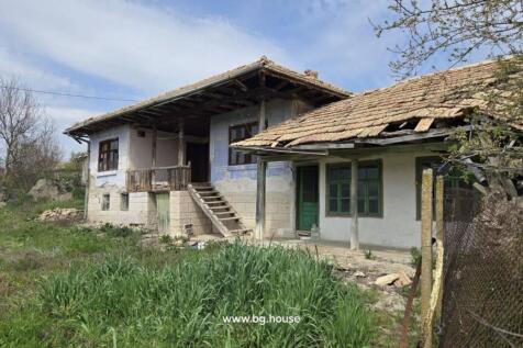 Side perspective of a Bulgarian house featuring stone basement, wooden balcony, tiled roof, and green doors and windows.